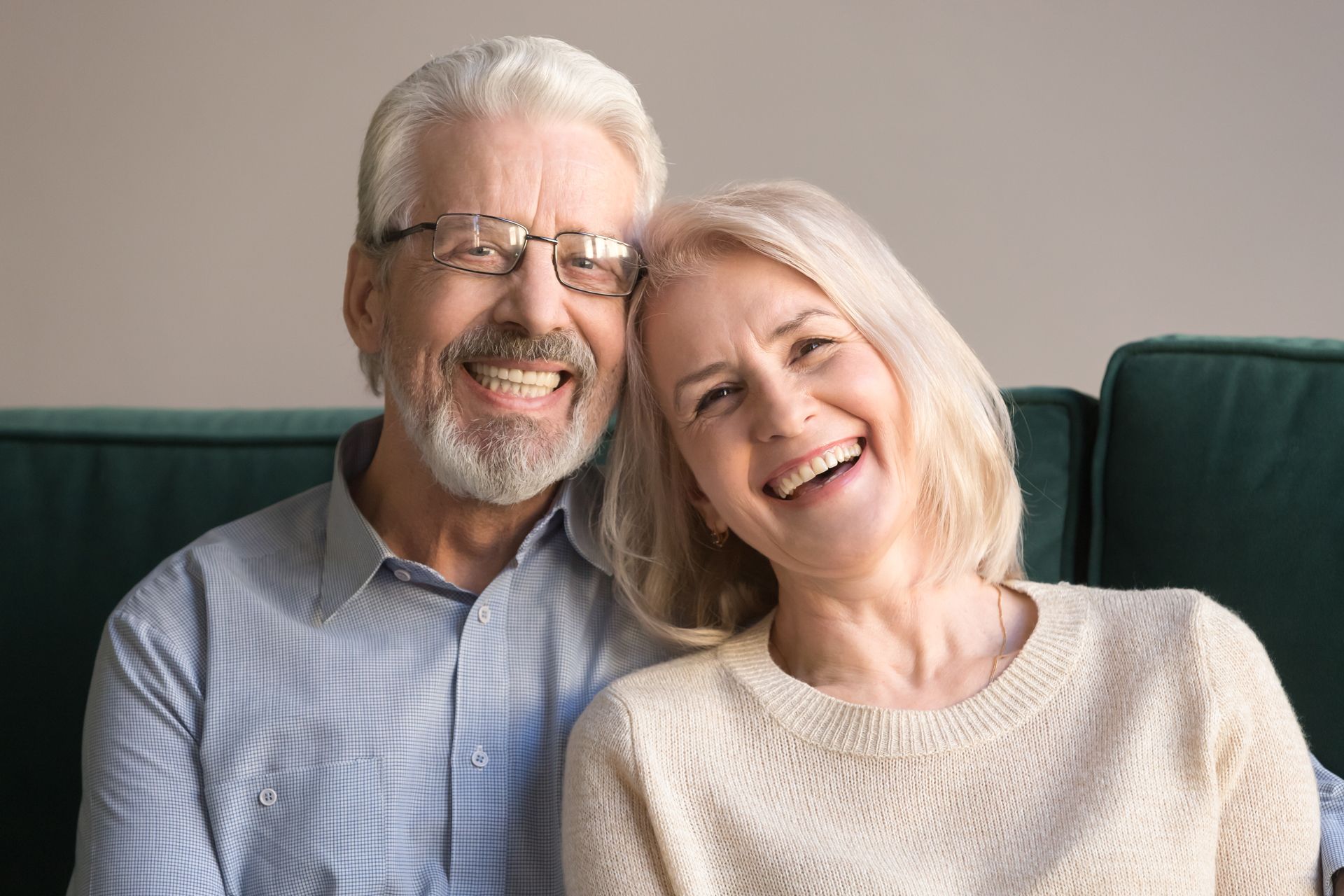 Two smiling people sitting close together on a sofa, with gray hair and a cozy indoor background.