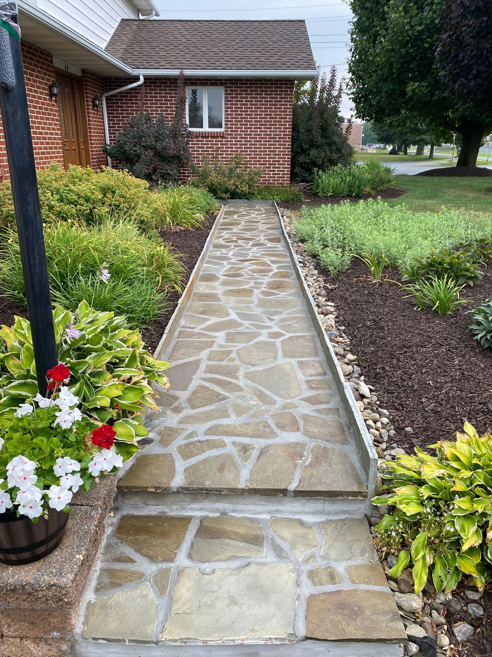 Stone walkway leading to a brick house, bordered by flowerbeds with green and brown plants.