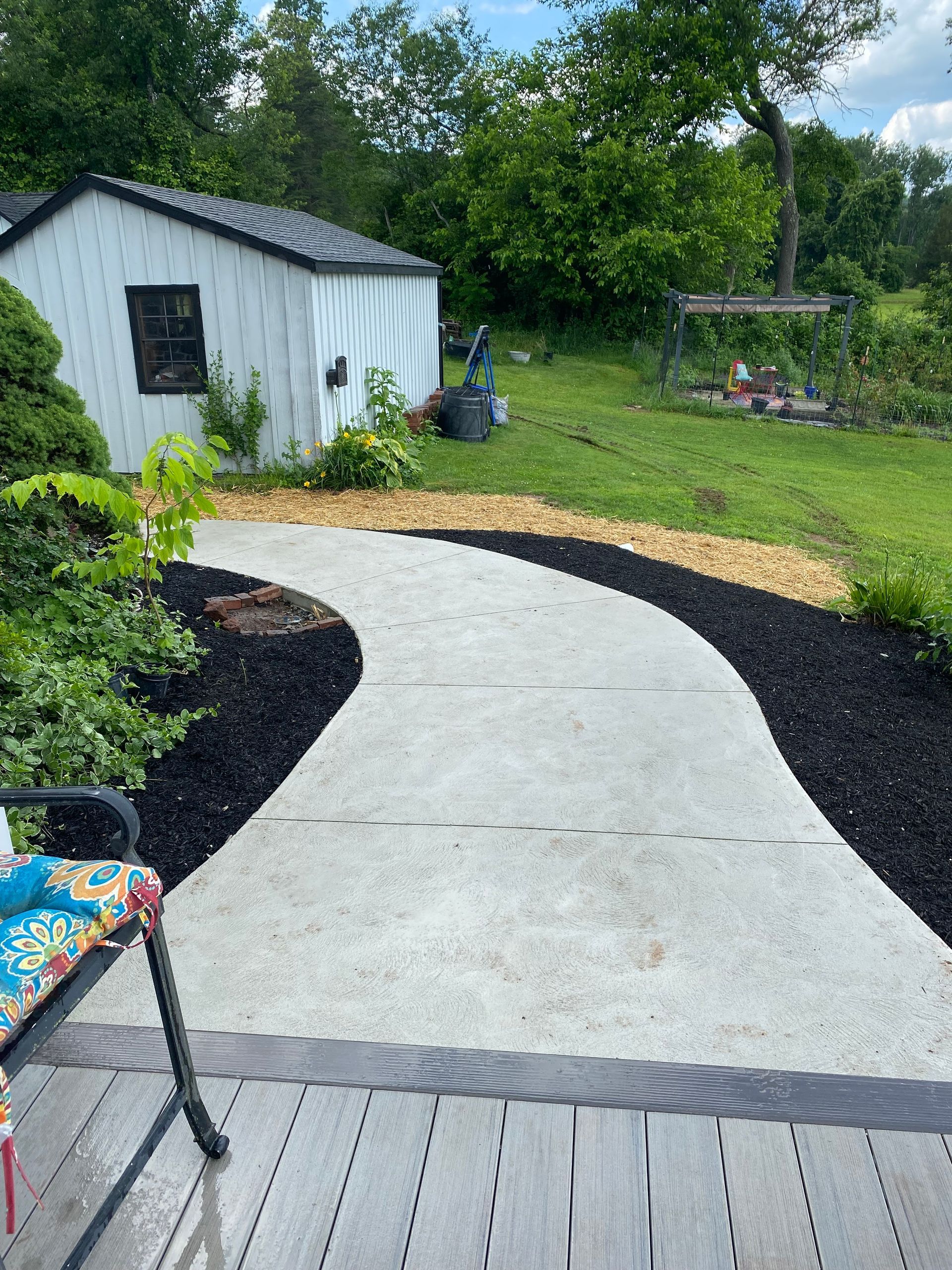Concrete pathway curves through a yard, edged by black and tan mulch, leading to a white shed.