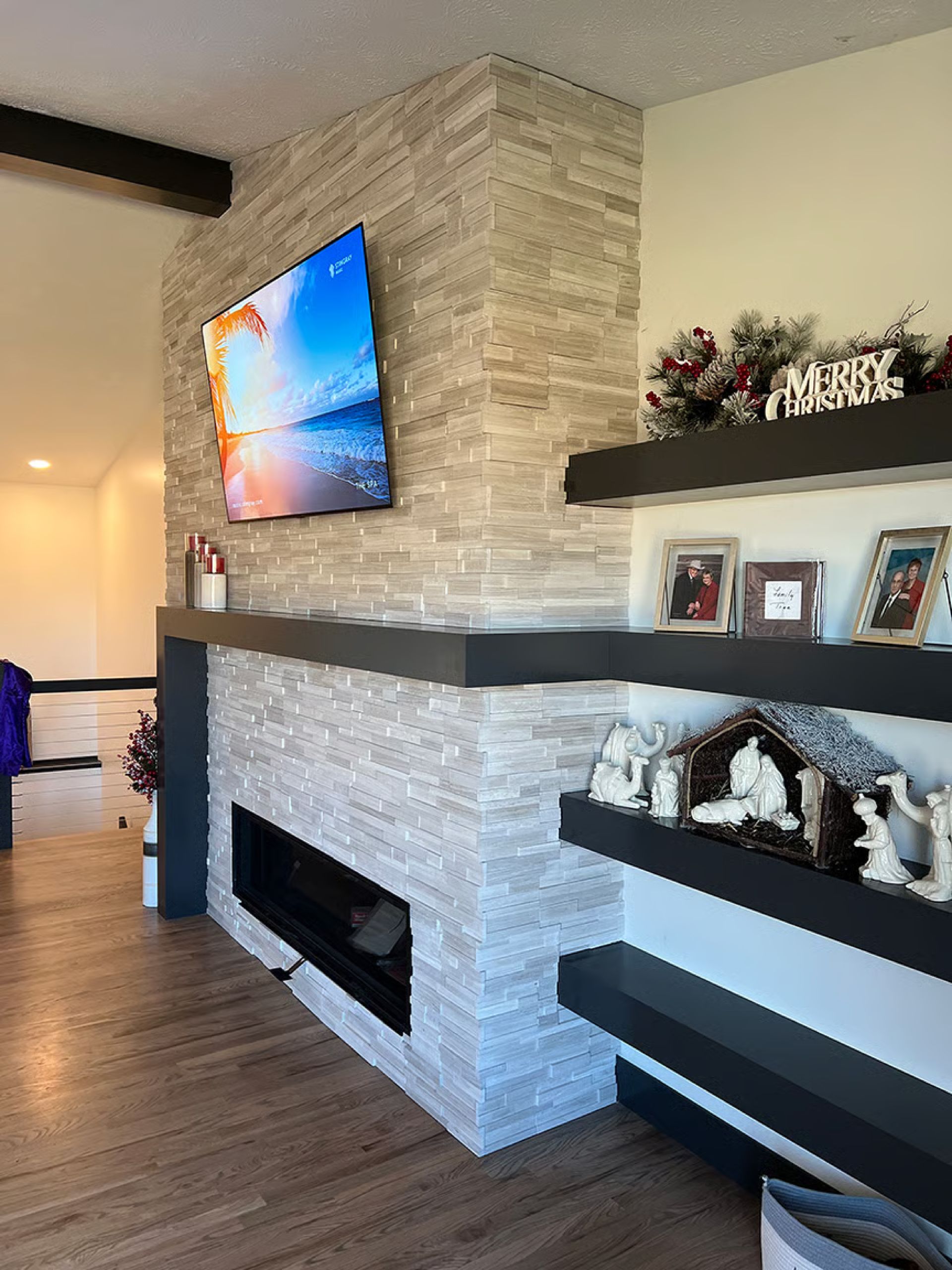 Fireplace with light stone facing, TV, black shelves with decorations, and wood flooring.