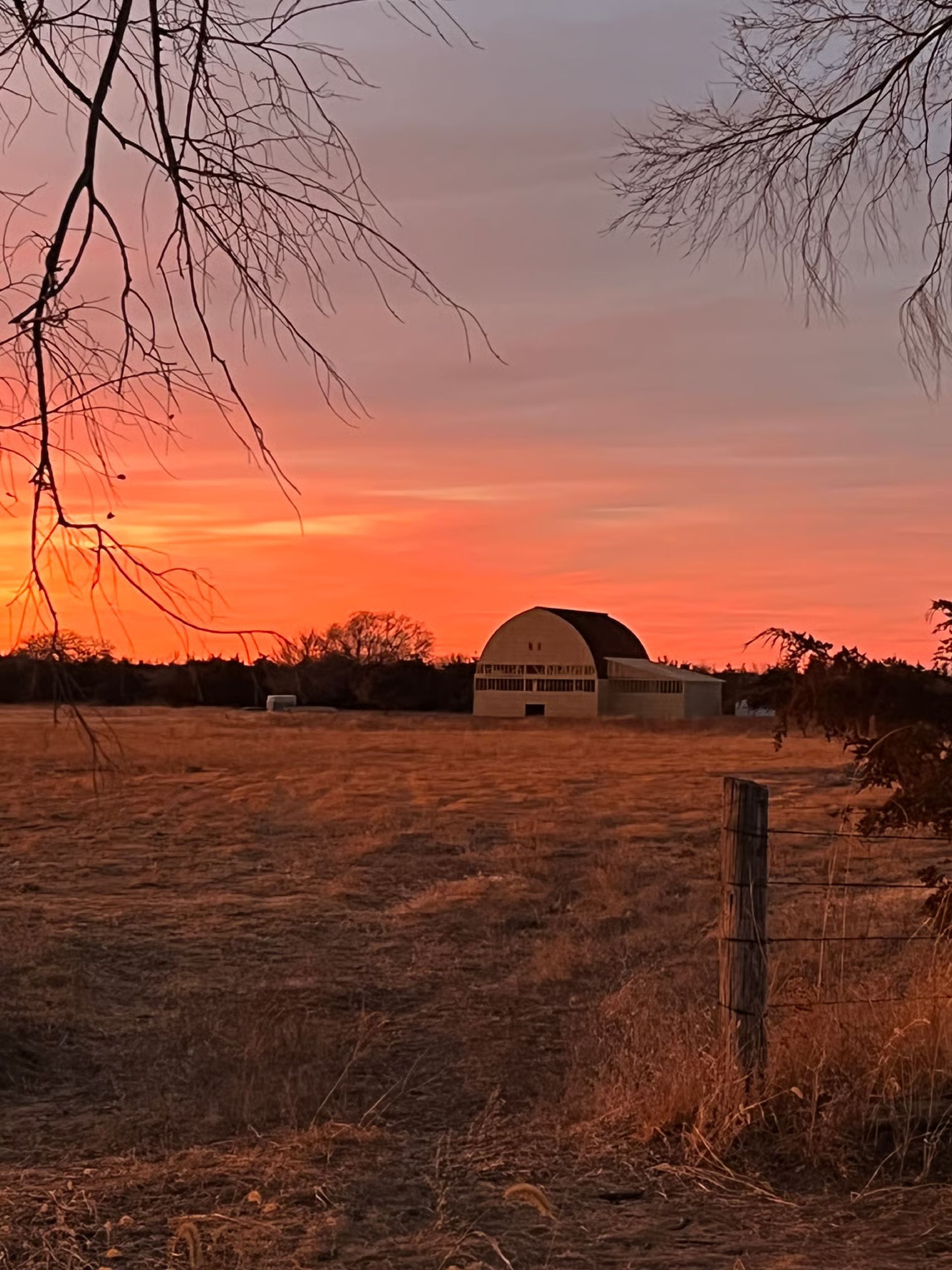 Sunset over a field with a barn and a fence post; orange and red sky.
