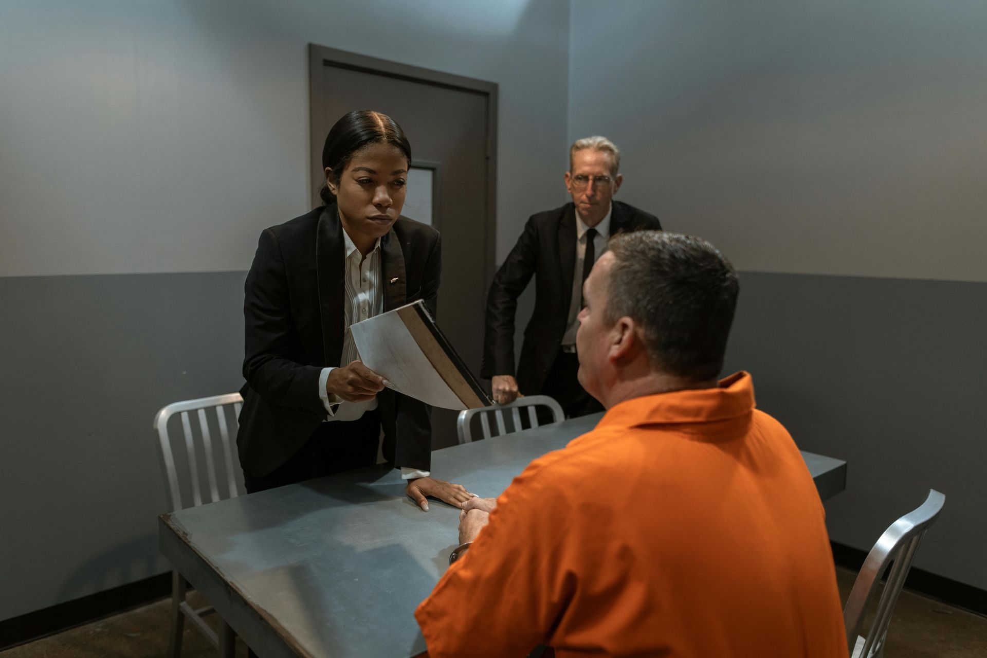 Police interrogation: a woman in a suit shows papers to an orange-clad man at a table, another man in a suit stands behind.