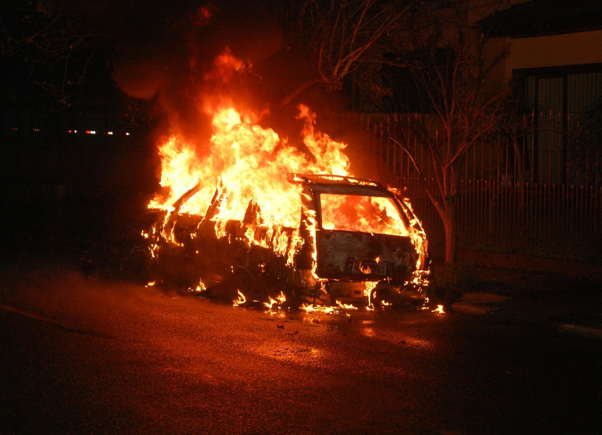 Car engulfed in flames at night, on a dark street, orange fire illuminating the surroundings.