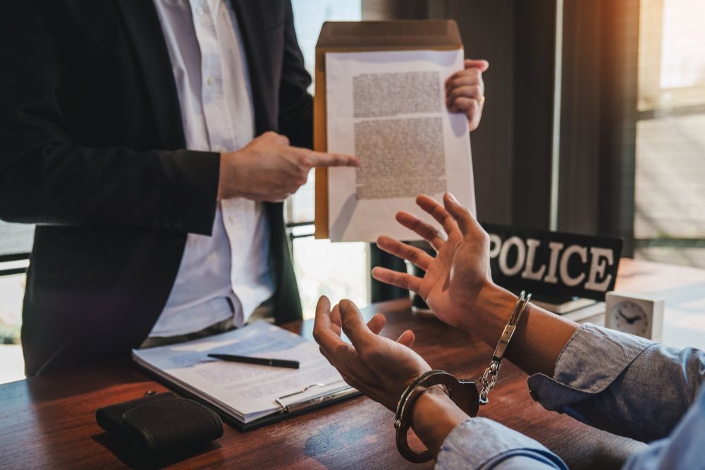 Police officer points at documents, handcuffed person in front, desk with