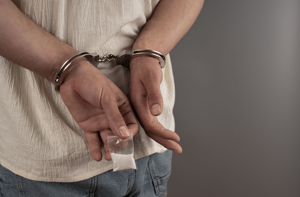 Person in handcuffs holding a small bag of white substance, likely drugs, behind their back.