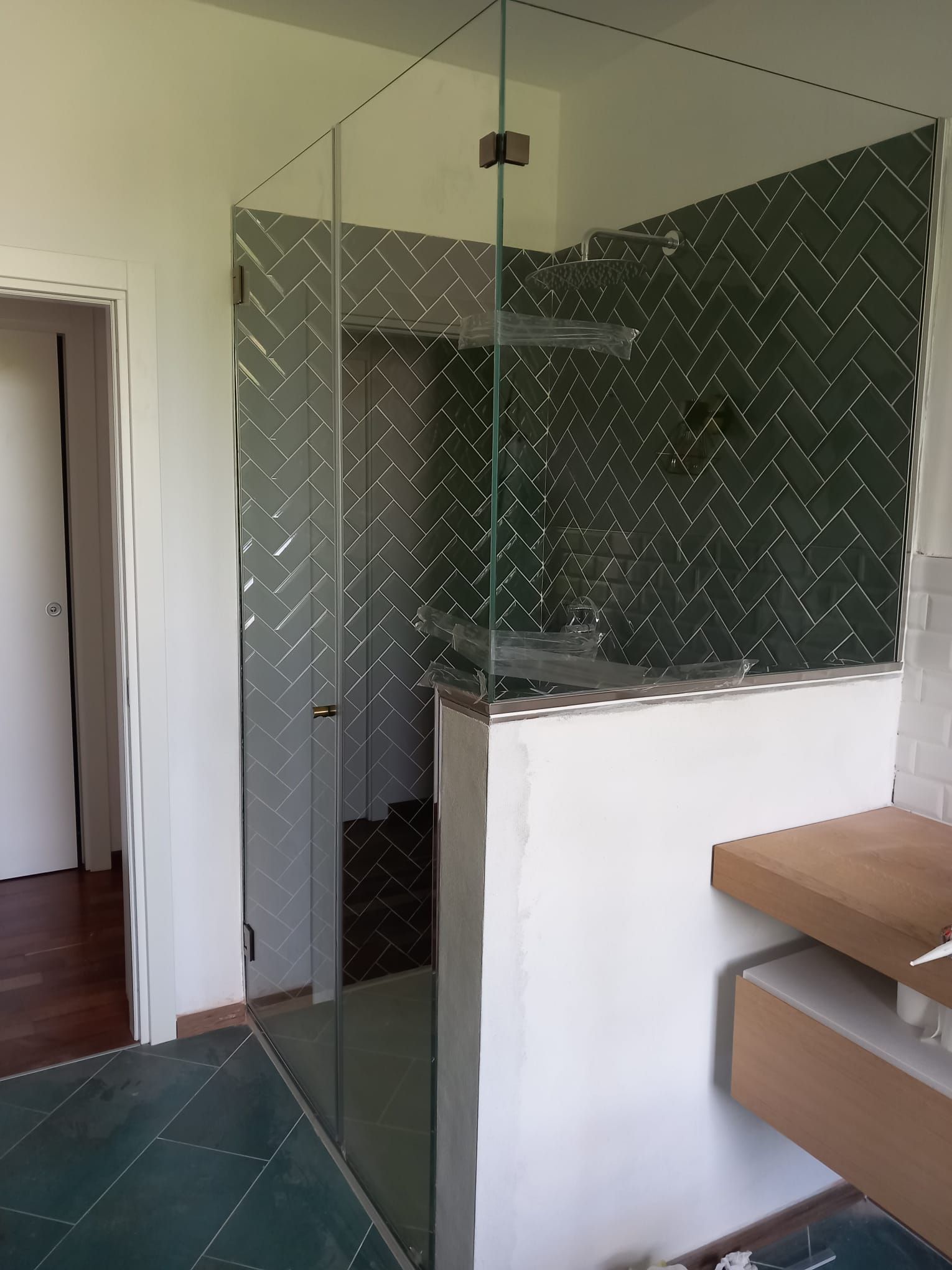 Bagno moderno con doccia piastrellata di verde, parete divisoria in vetro e panca in legno accanto a un muretto bianco.