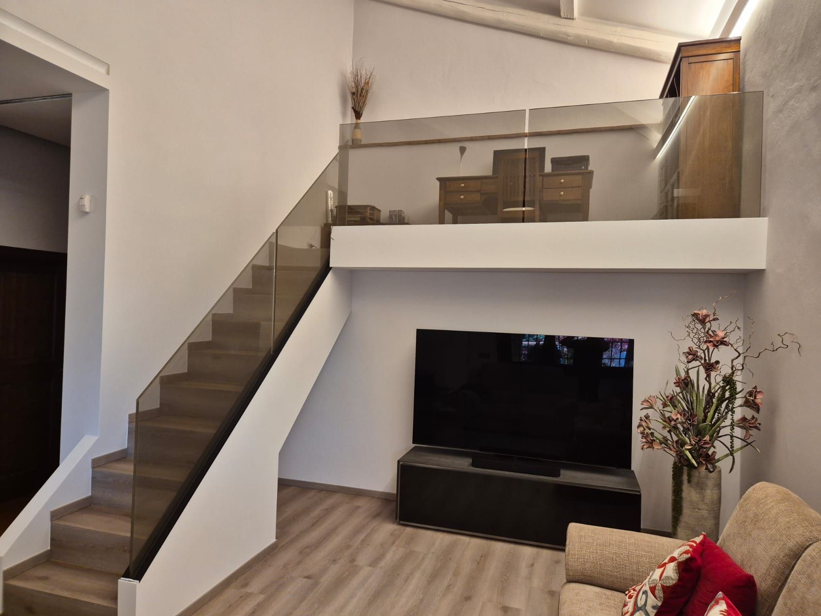 Soggiorno con scala, ringhiera del soppalco, TV e divano beige in una luminosa casa moderna.