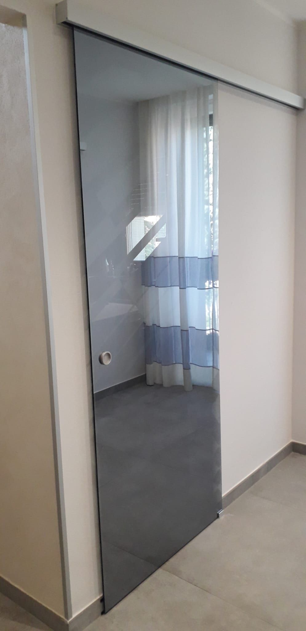La porta a specchio di un armadio nel corridoio riflette una camera da letto con un letto blu e bianco e tende trasparenti.