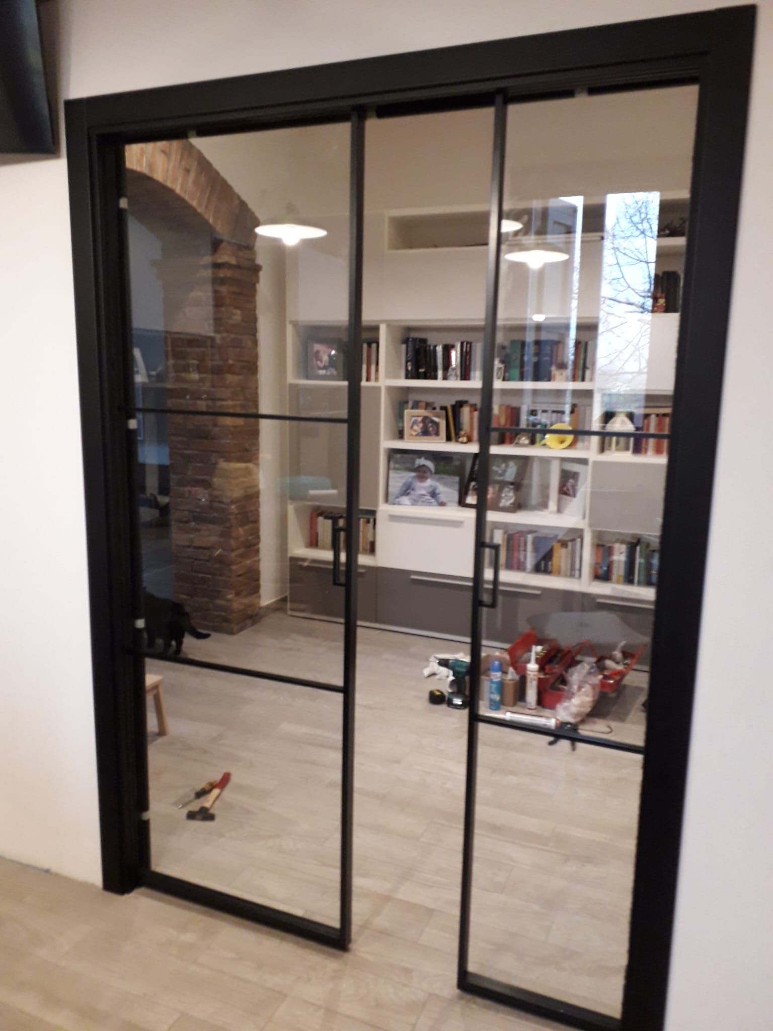 Le porte a vetri con cornice nera dell'ufficio si aprono su una stanza con scaffali, libri e materiale scolastico su un pavimento chiaro.