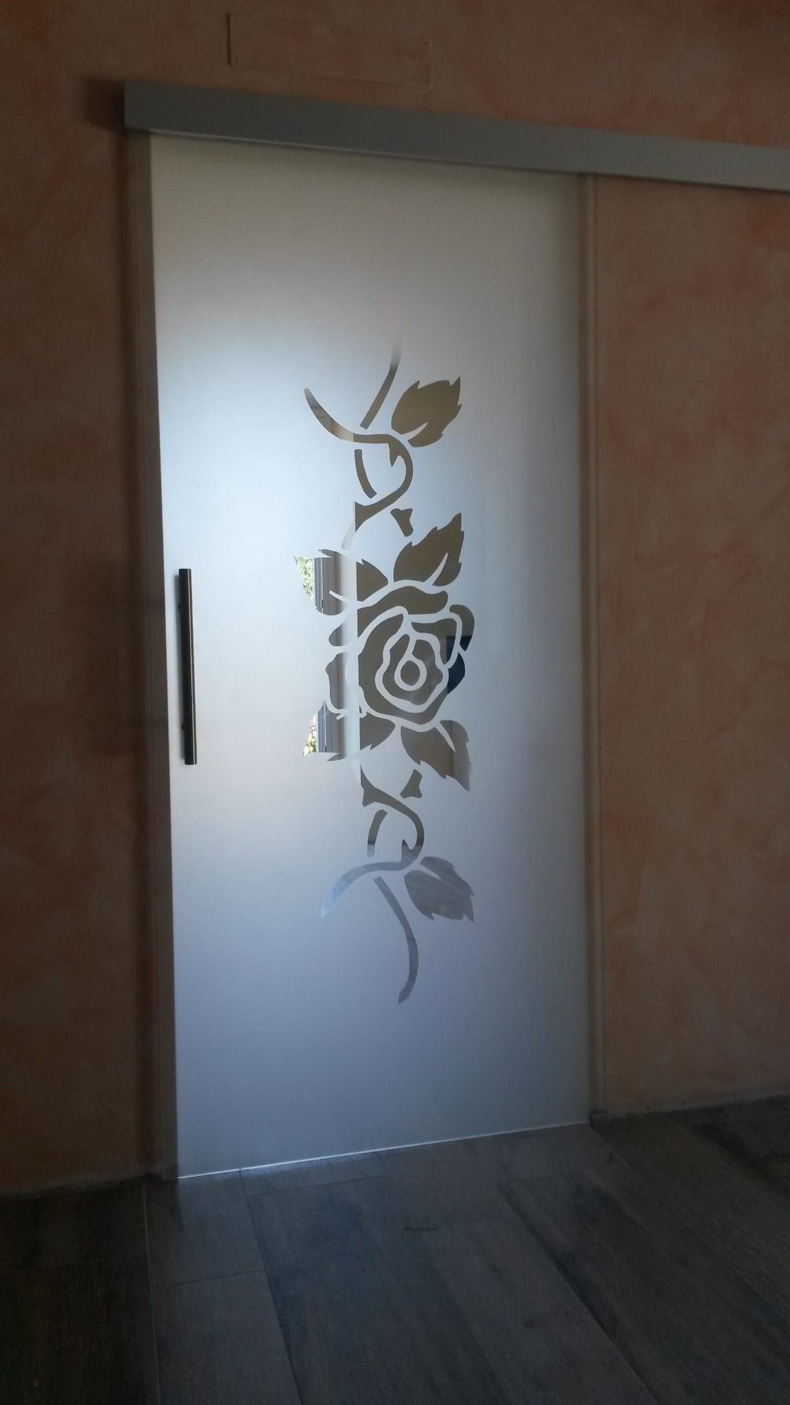 Porta in vetro satinato con motivo floreale verticale e maniglia scura in un corridoio poco illuminato.