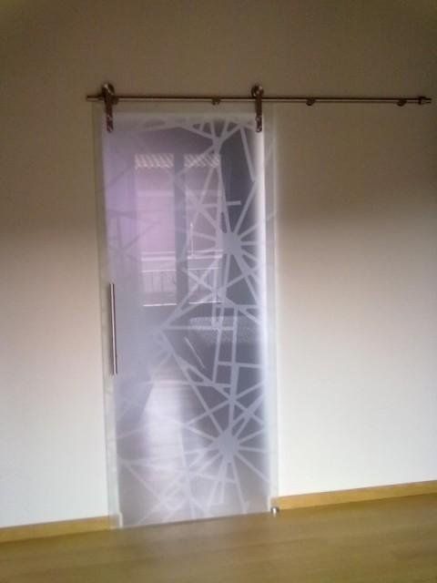 Una tenda bianca trasparente con motivo geometrico pende da una porta, accanto a una parete bianca e un pavimento in legno.