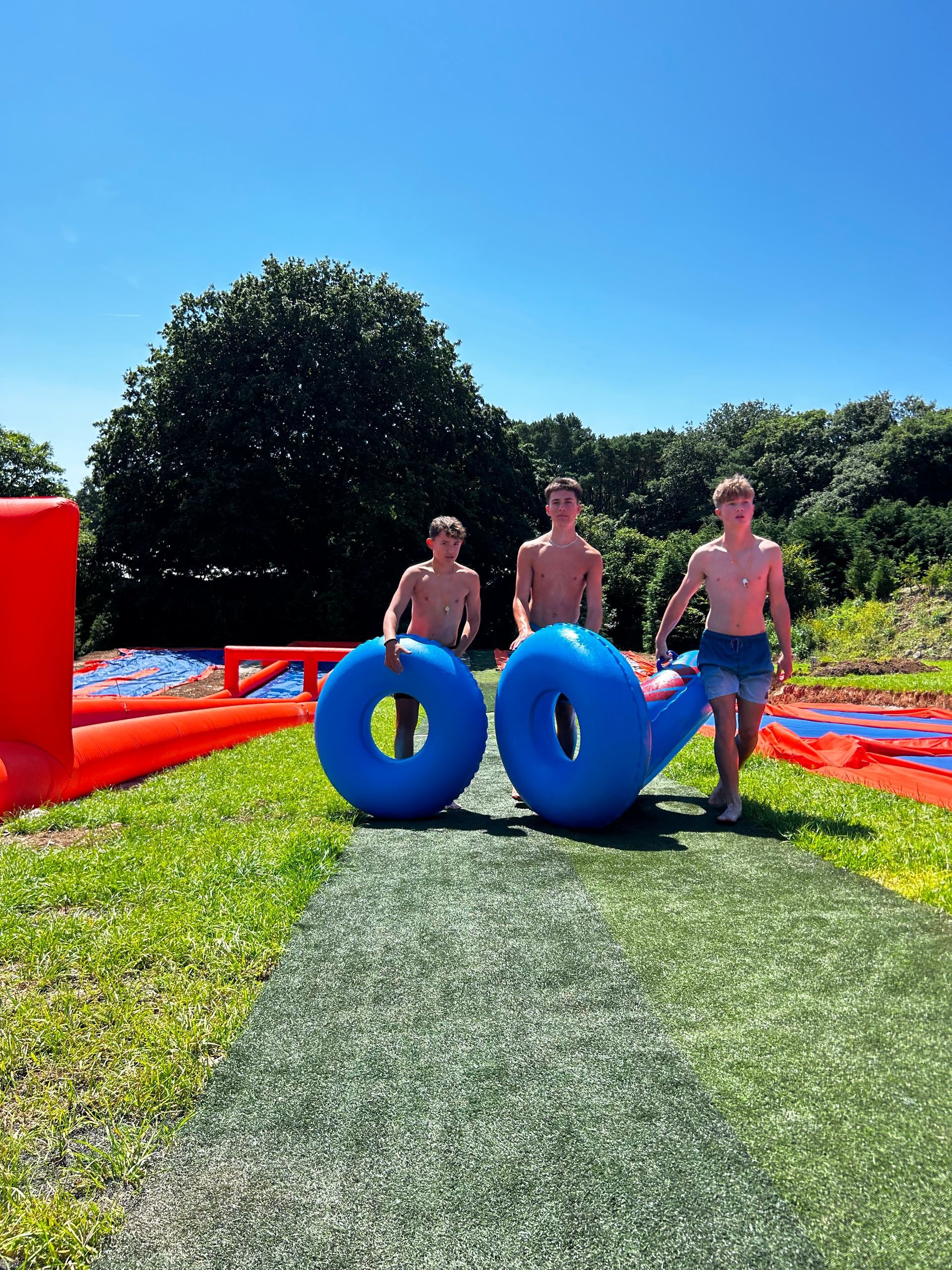 Three shirtless individuals carry blue inflatable rings across a sunny, grassy path near outdoor water play equipment.