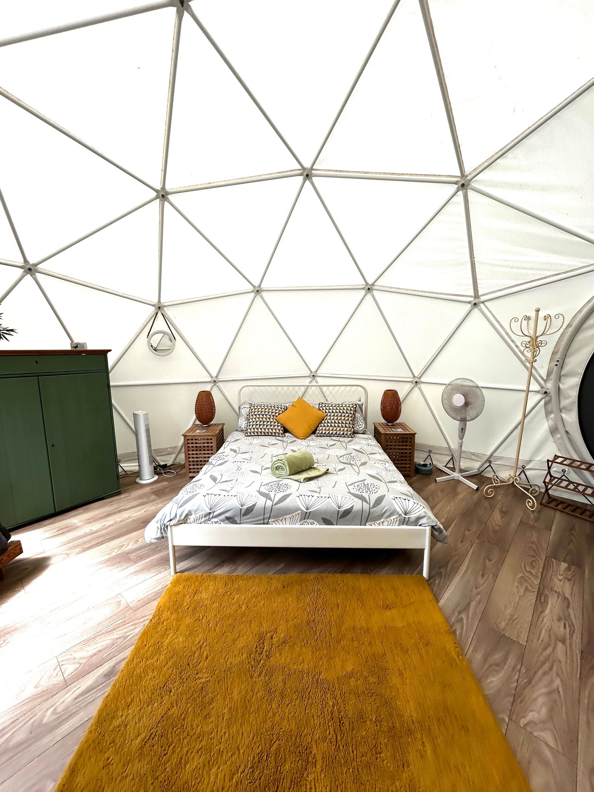 A geodome tent interior featuring a central bed with a mustard yellow pillow, a yellow rug, and a green storage unit.