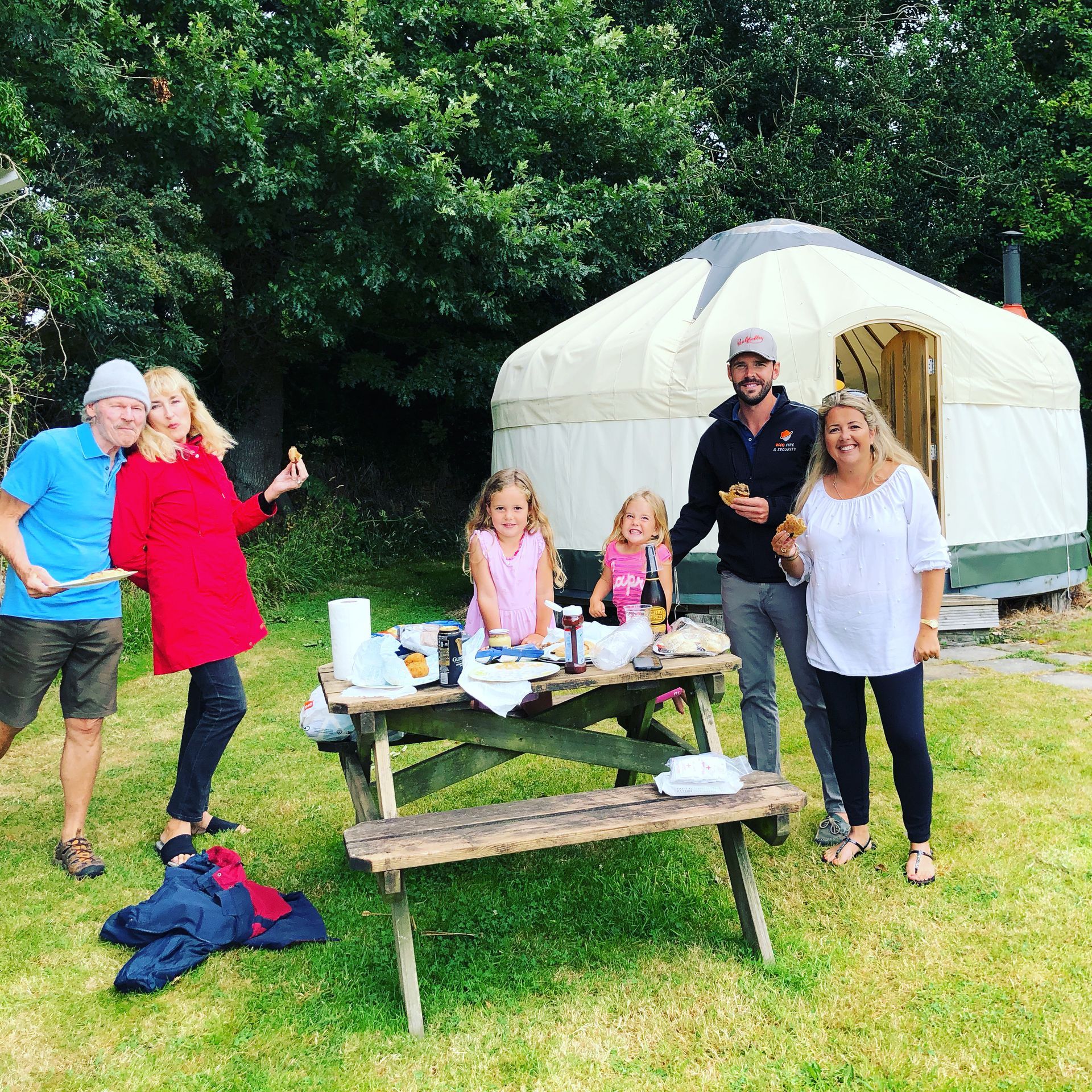 A group of six people posing for a photo around a wooden picnic table in front of a white yurt on a grassy field.