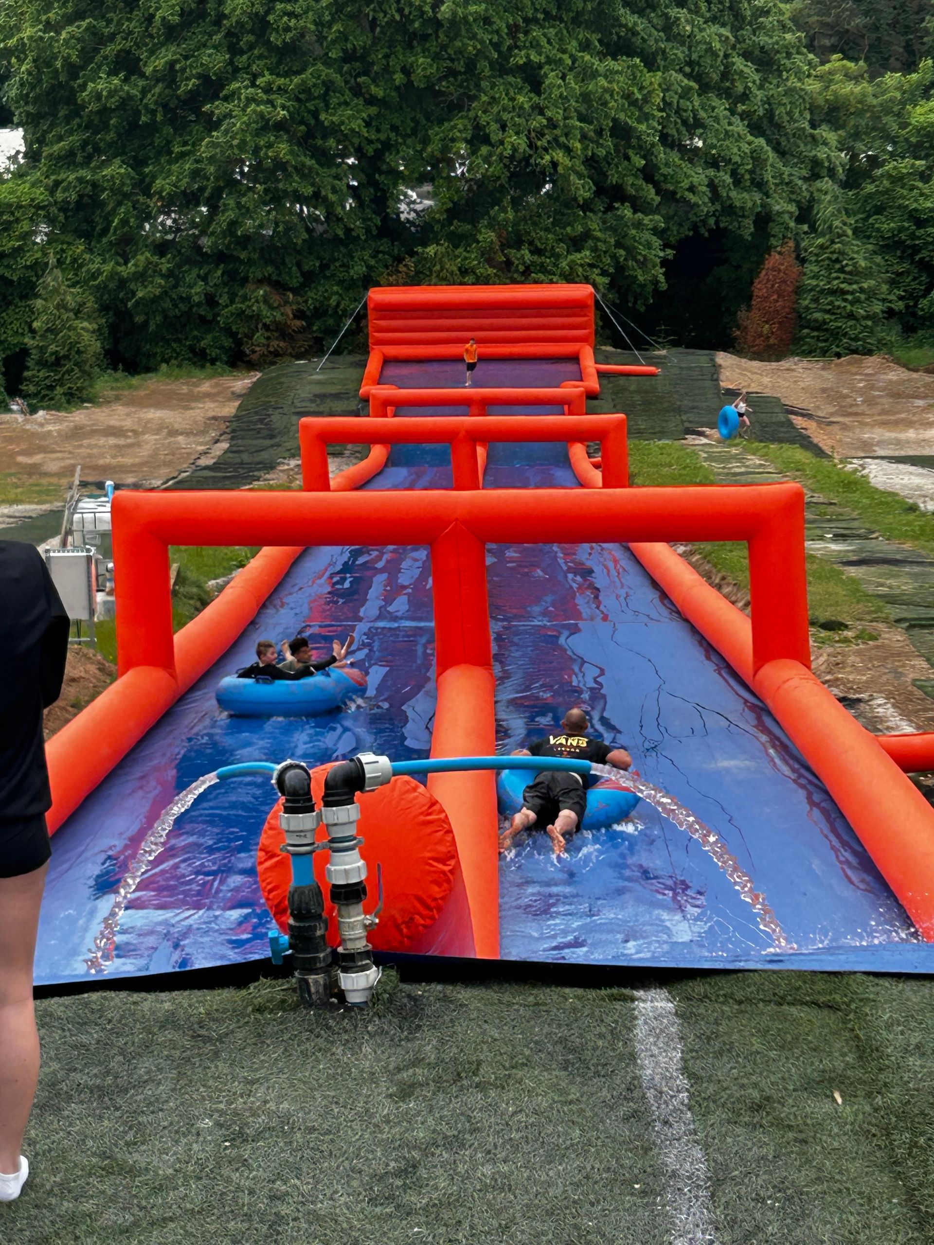 Two people on inner tubes slide down a dual-lane inflatable water slide on a grassy outdoor field.