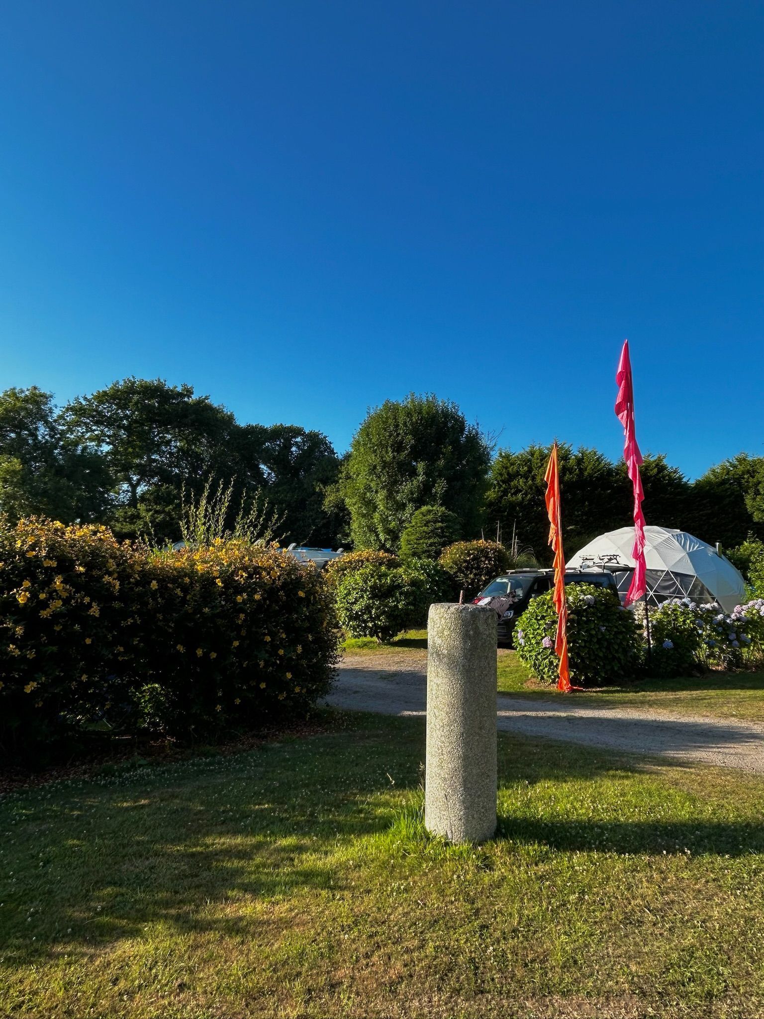 A bright, sunny campsite with a stone pillar in the foreground, a trimmed hedge, trees, and a white dome tent behind.
