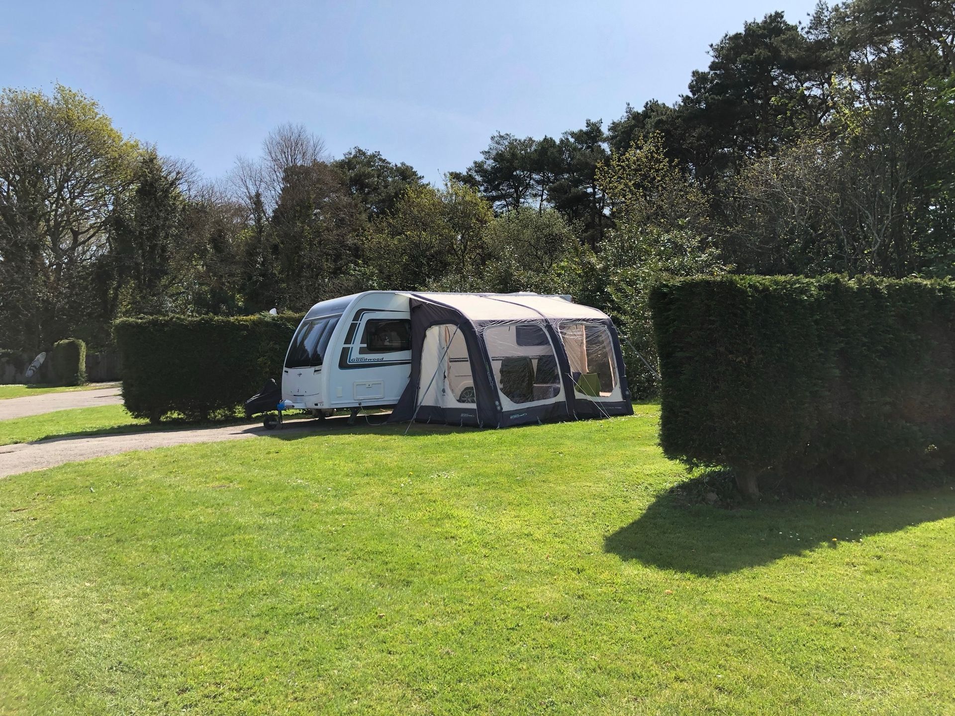 A white travel trailer with a large inflatable awning parked on a grassy pitch between two tall green hedges.