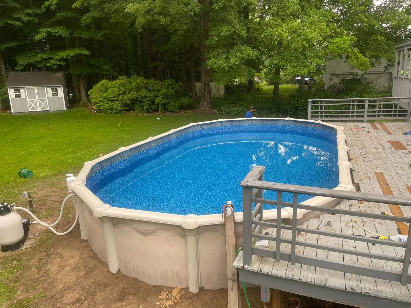 Above-ground backyard pool with blue water beside a wooden deck and green lawn, surrounded by trees