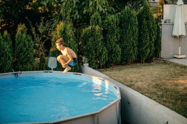 Person crouching by a backyard pool beside tall hedges and a patio umbrella