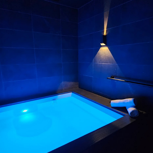Woman in black bikini seated cross-legged in a sauna, with blue and amber lighting.