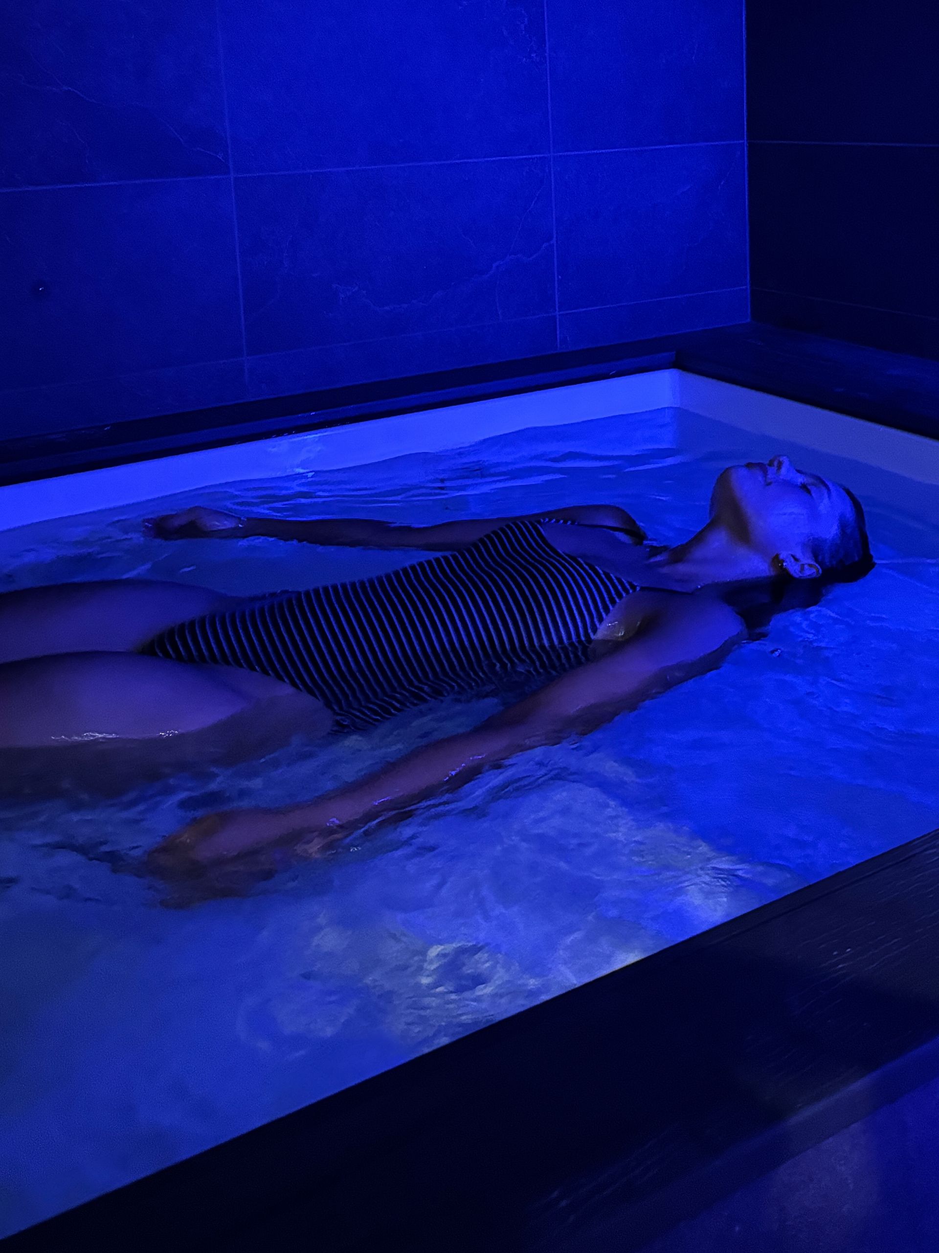 Woman in black bikini seated cross-legged in a sauna, with blue and amber lighting.