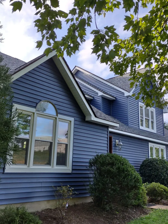 Blue house exterior with white-trimmed windows, steep roof, and leafy tree branches overhead