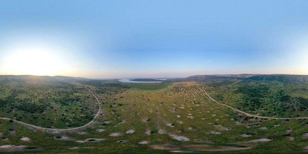 Aerial panoramic drone view of Akagera National Park at sunrise showing open savannah game drive track acacia woodland and Lake Ihema between sandstone ridges eastern Rwanda