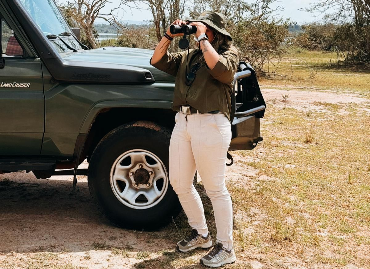 A guest in safari attire using binoculars beside a 4x4 safari vehicle, with a lake visible in the background.