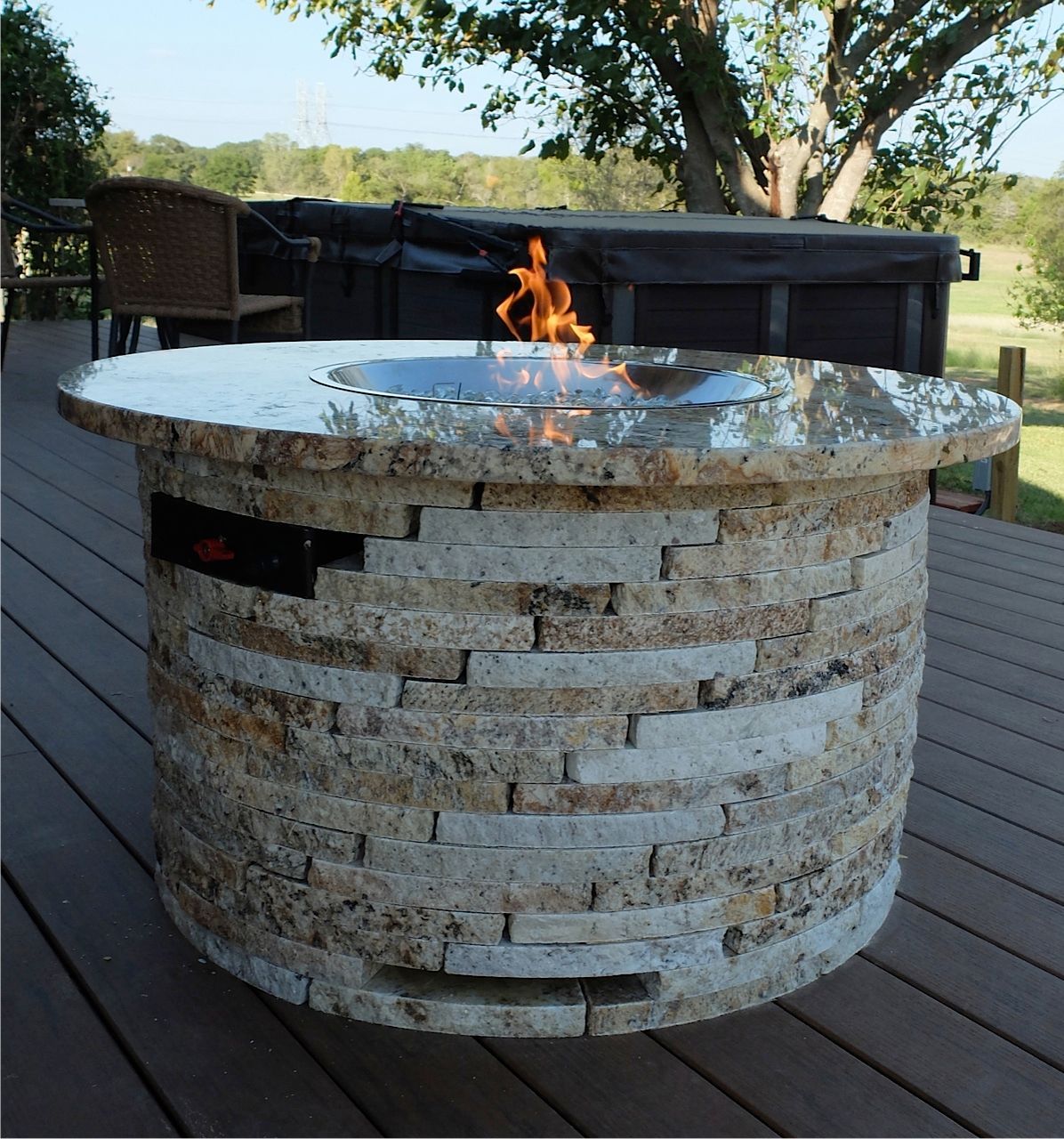 Stone fire pit with a small flame on a wooden deck, outdoors with a couch and trees in the background