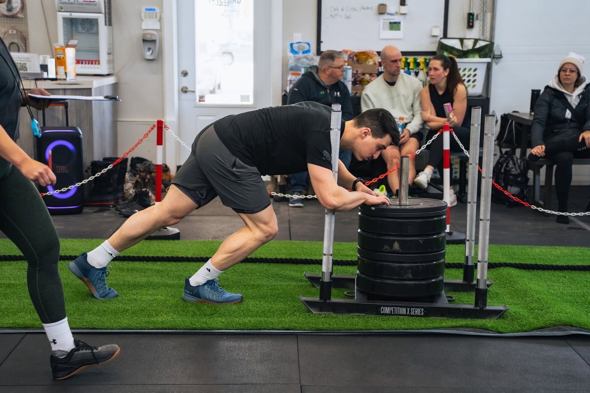 Man pushing a loaded sled in a gym, leaning forward on turf while spectators watch behind a rope barrier