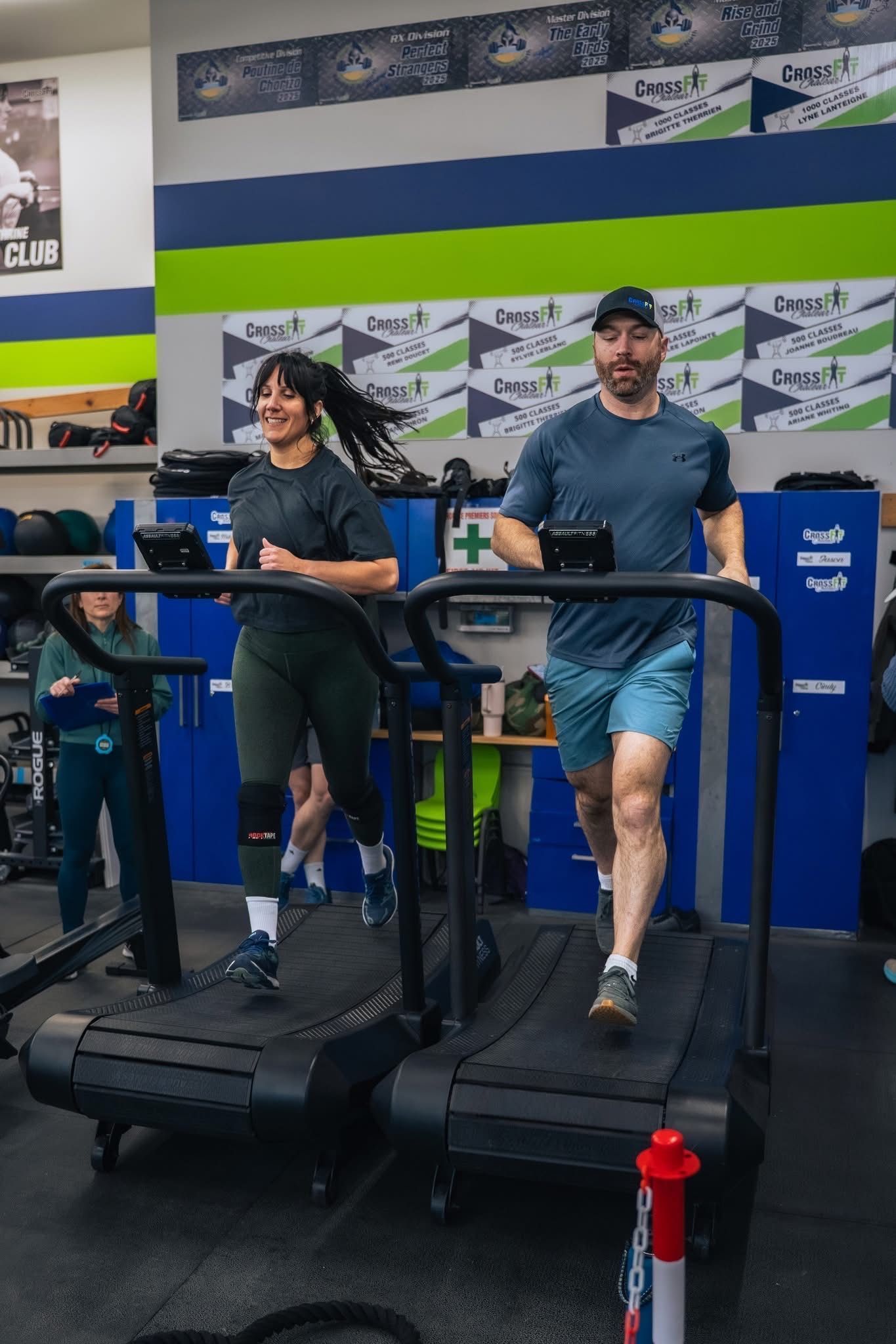Two people walking on treadmills in a gym, with workout equipment and branded wall banners behind them.