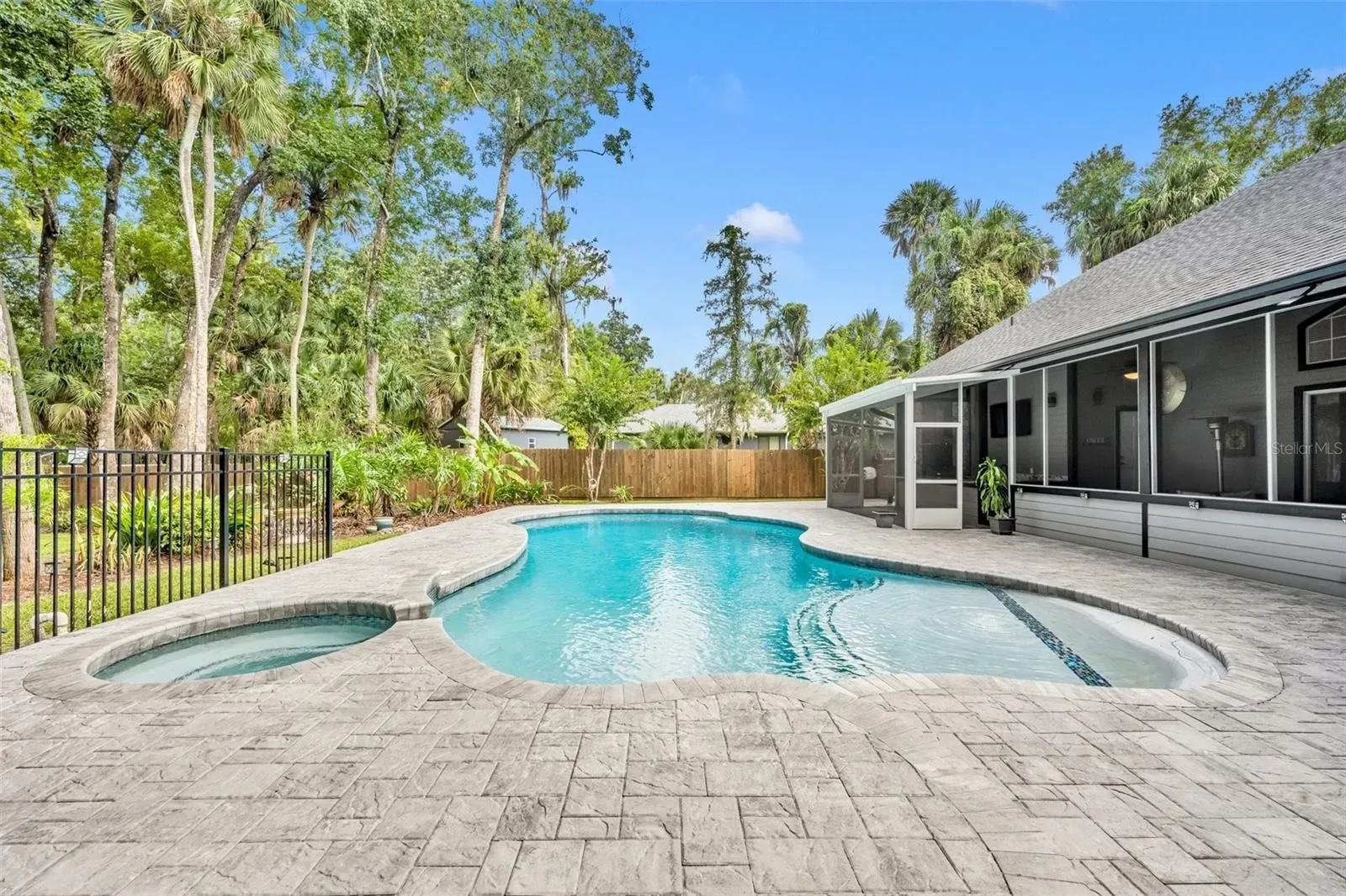 Backyard pool with stone deck, screened patio, and trees beyond a black fence