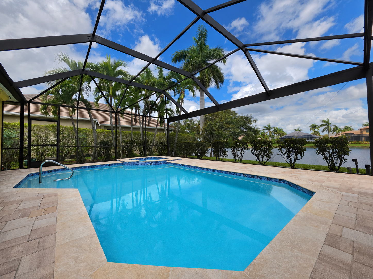 Screened lanai with a blue pool, tan pavers, palm trees, and a lake view under a cloudy sky