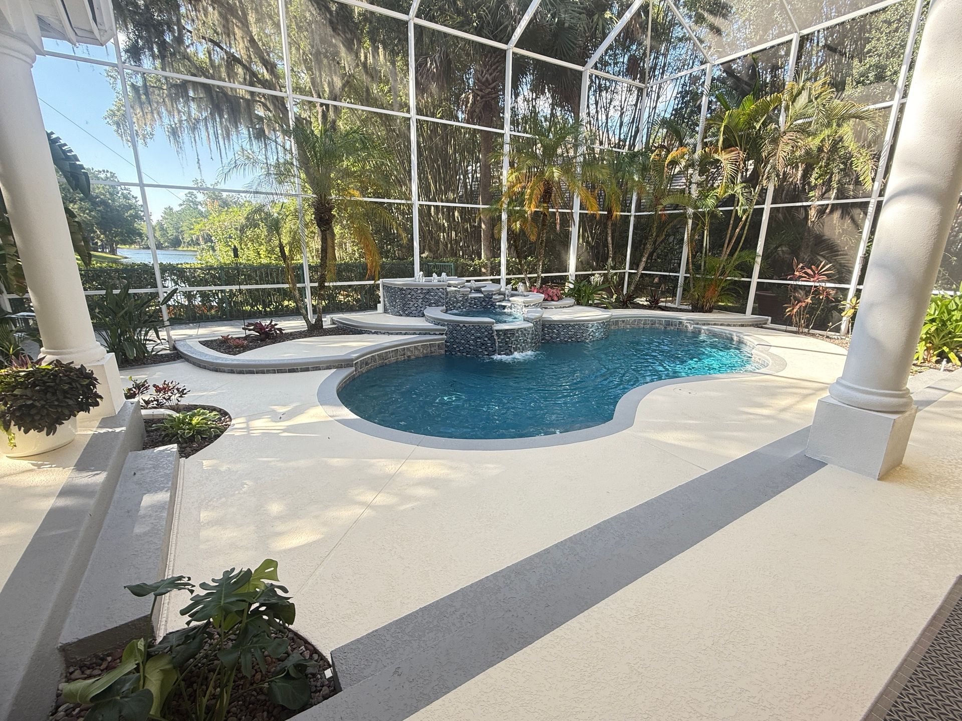 Screened patio with a curved blue pool, white columns, and tropical plants in bright sunlight