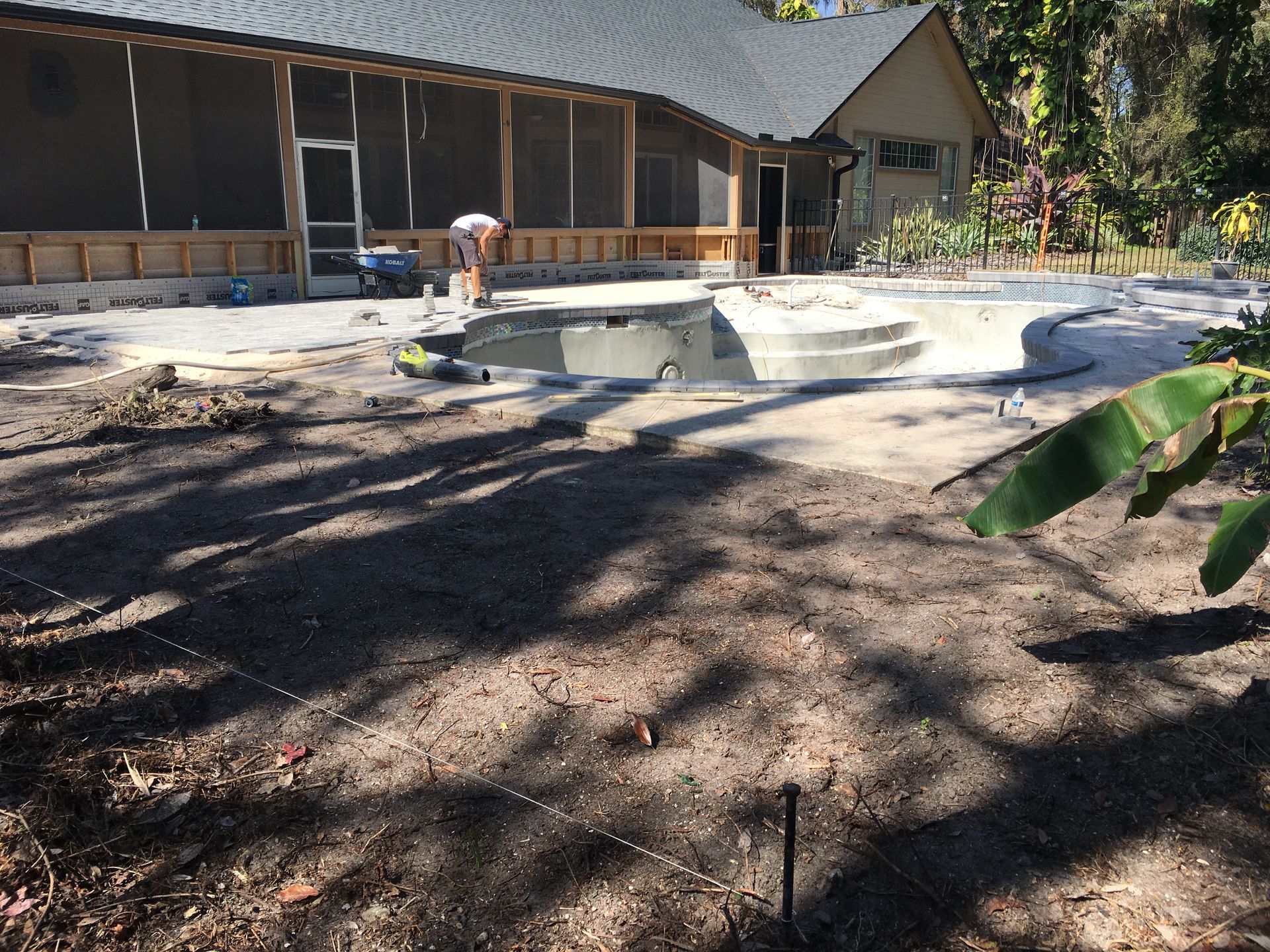 Partially built concrete pool beside a house, with a person working nearby under trees