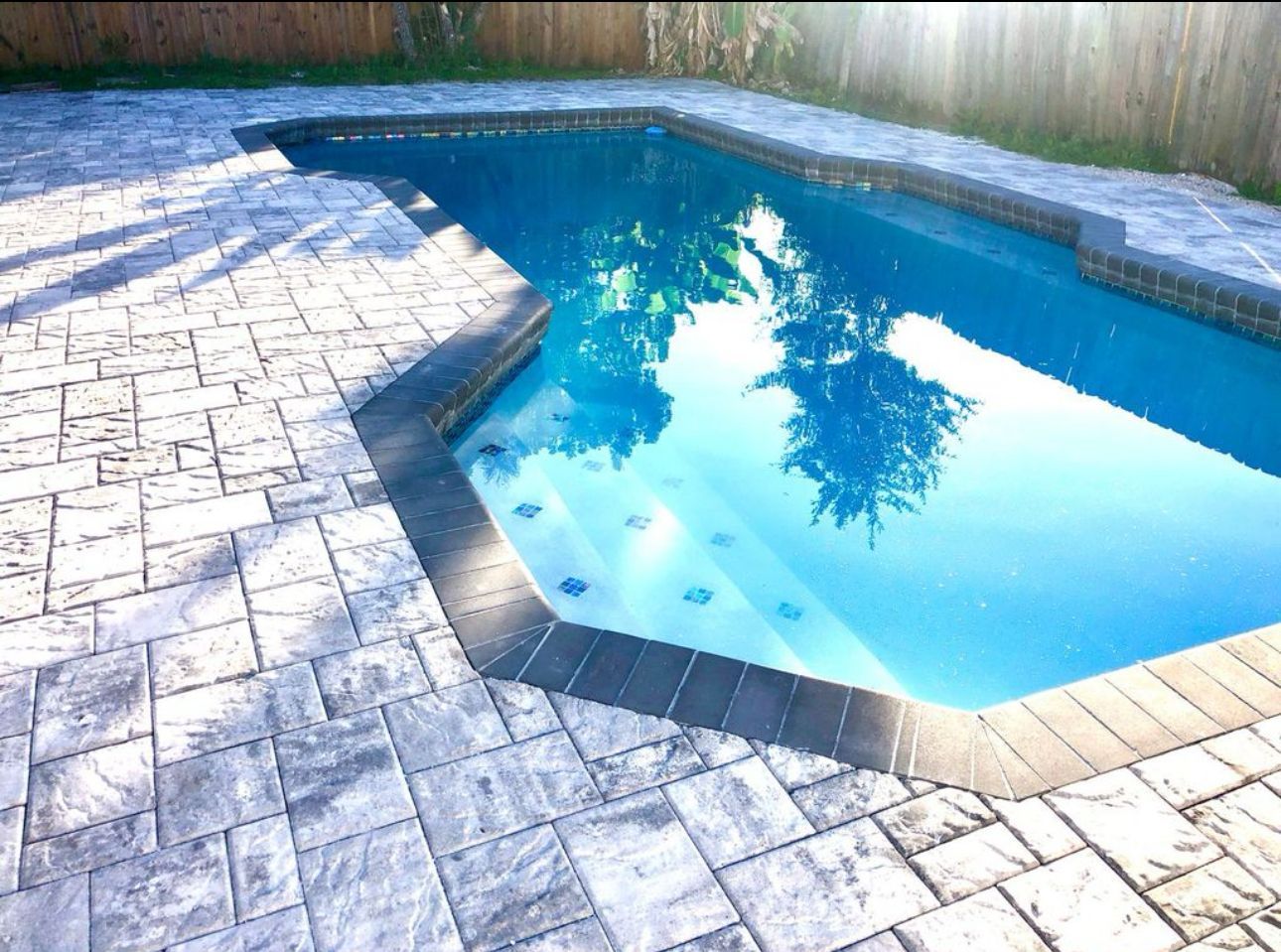 Backyard swimming pool with blue water, stone patio, and wooden fence in bright sunlight