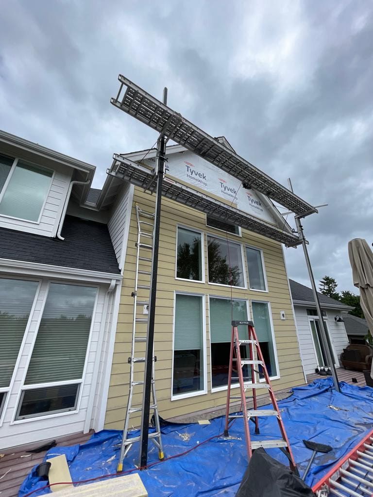House exterior under construction, scaffolding, ladders, and blue tarp. Overcast sky.