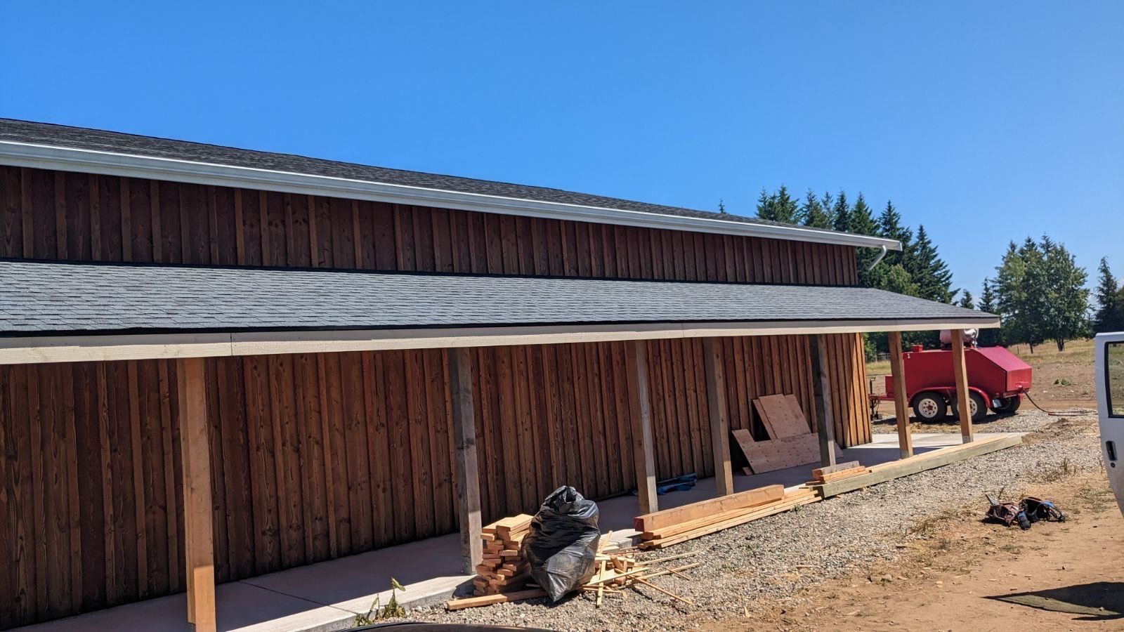 Newly built brown wooden building with gray roof, porch supported by wooden posts, under blue sky.