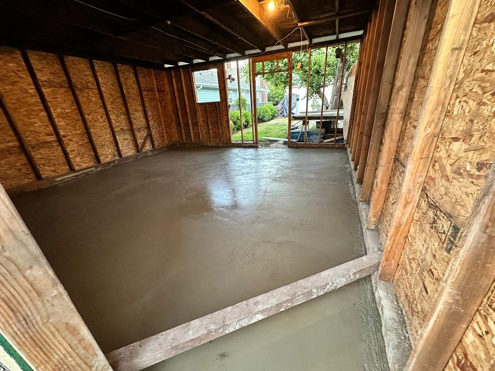 Newly poured concrete floor inside a wood-framed structure, with an open doorway leading outside.