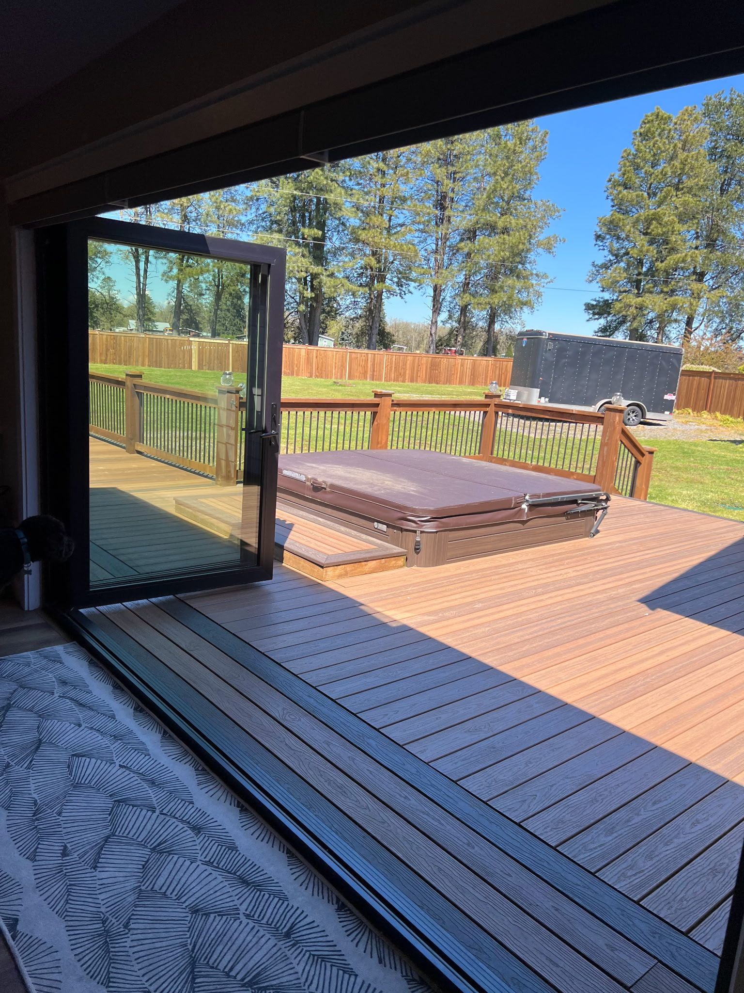 Open glass doors lead to recently built wooden deck with a hot tub, a lawn, and a fence under a blue sky.