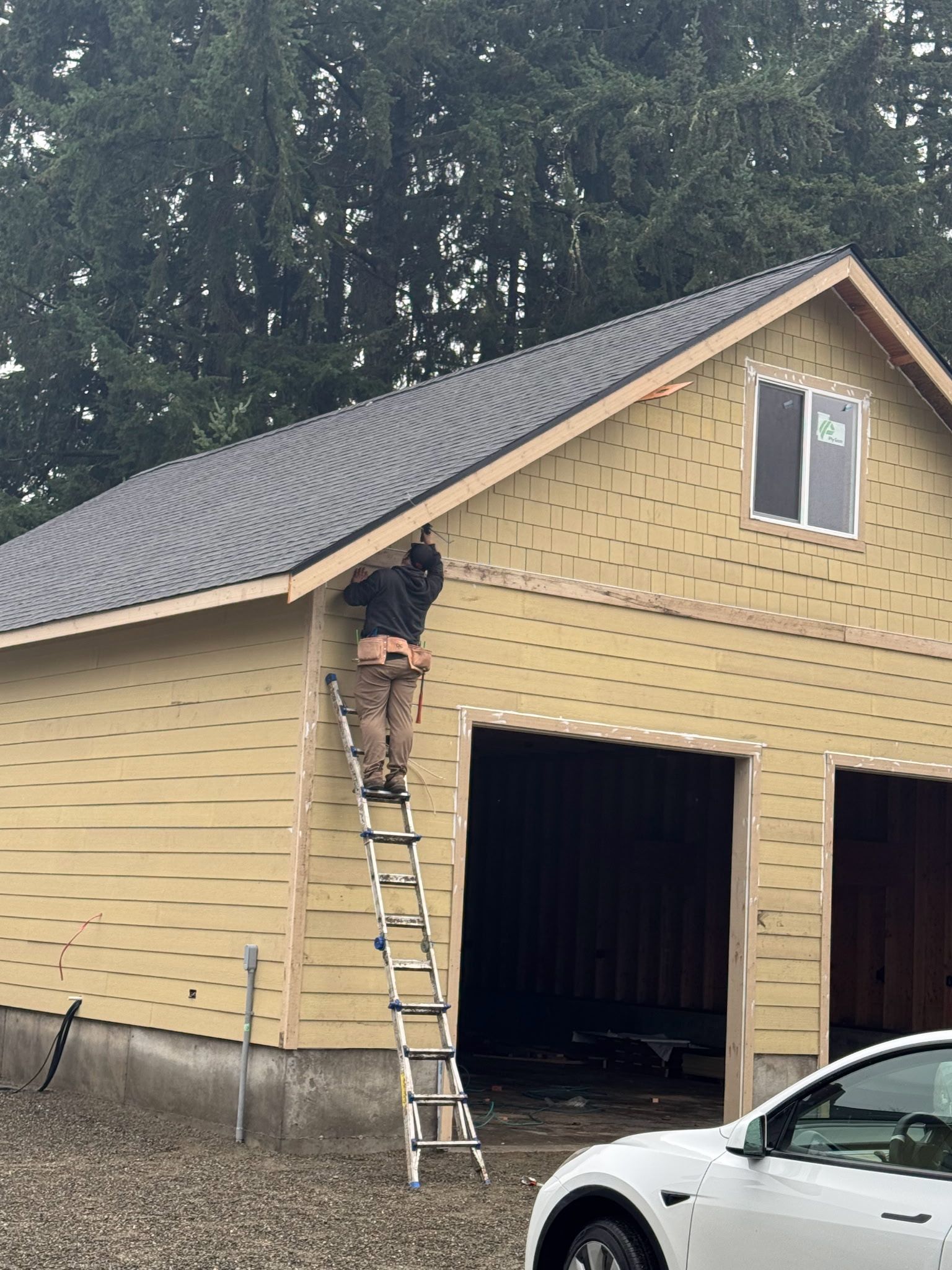 Harmon & Harmon Construction on a ladder working on the roof of a garage with yellow siding and a white car parked in front.