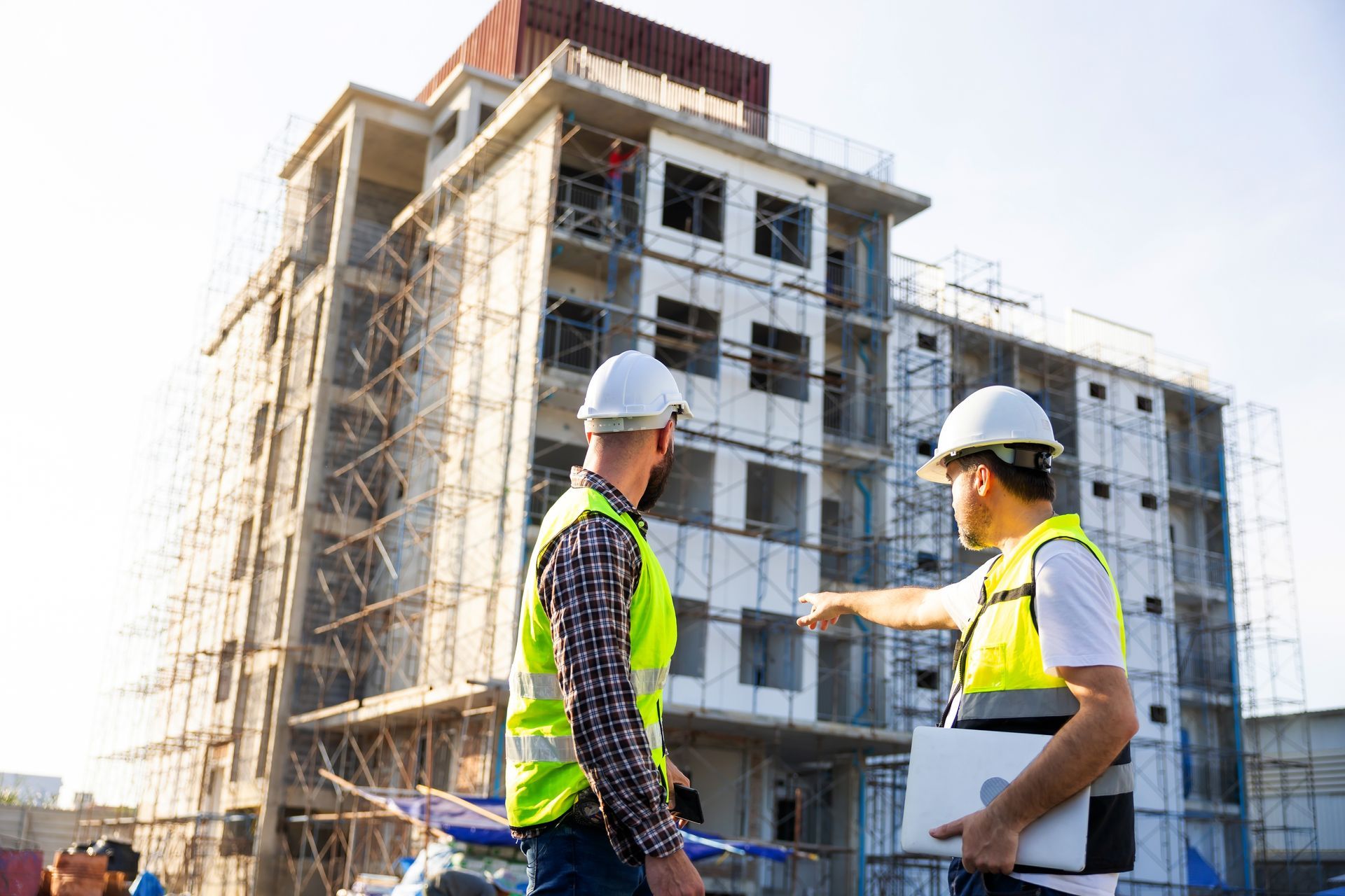 Two construction workers in safety vests and hard hats stand before a building under construction.