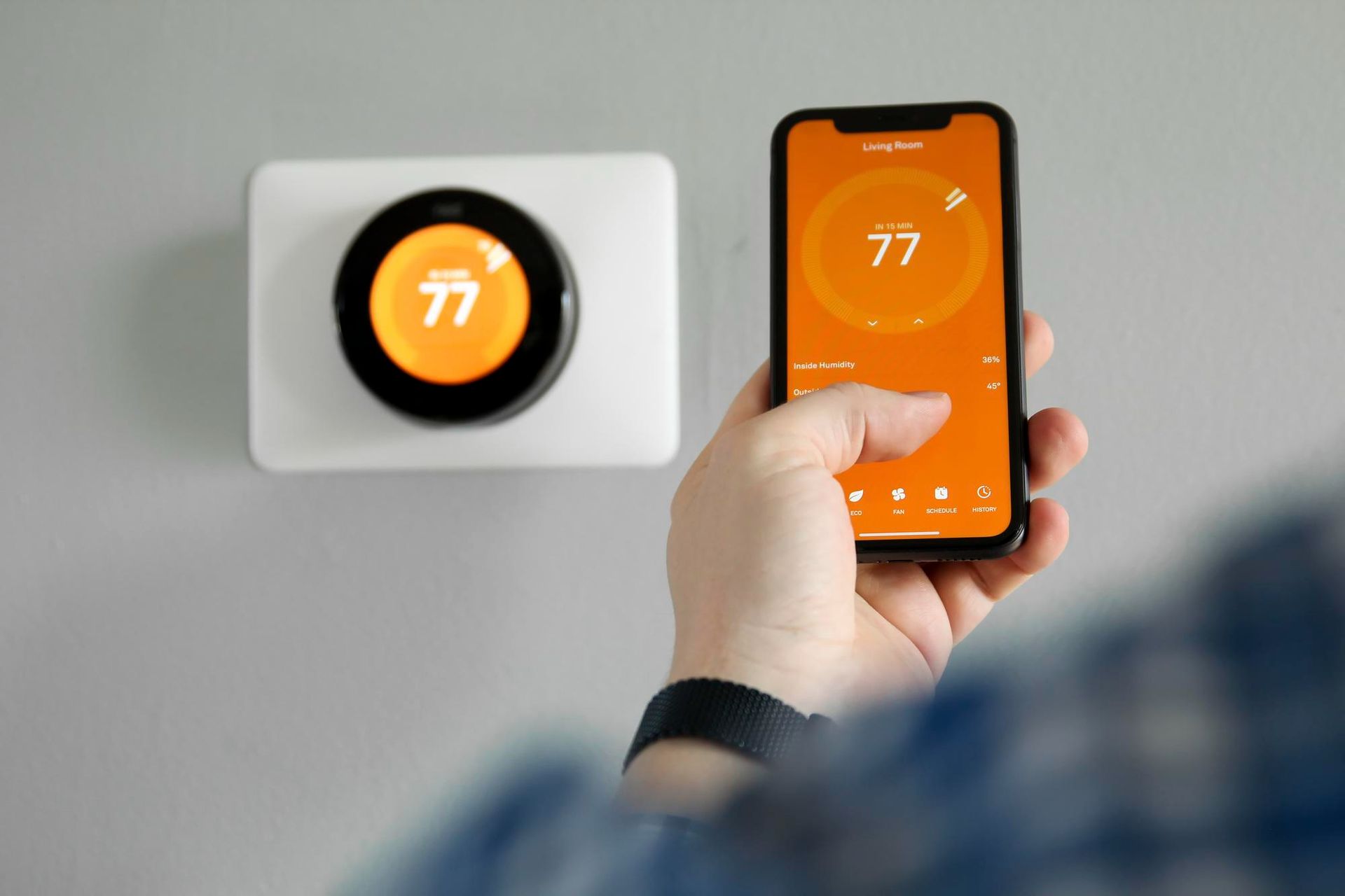 Hand holding a smartphone controlling a wall thermostat showing 77° on an orange screen