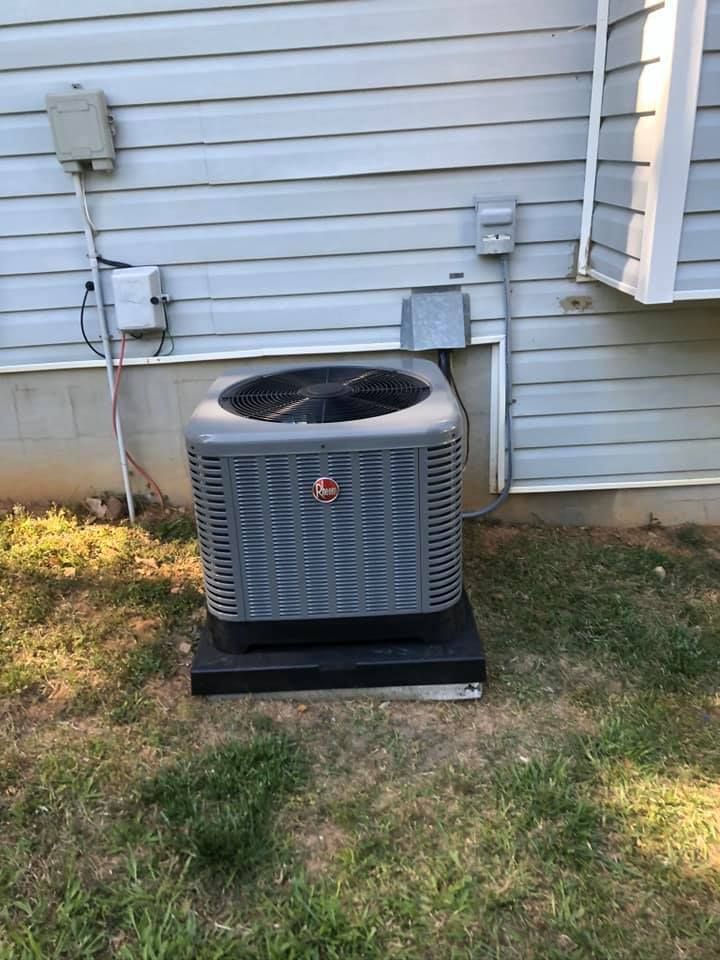 Outdoor air conditioner unit beside a house with white siding and electrical boxes on the wall.