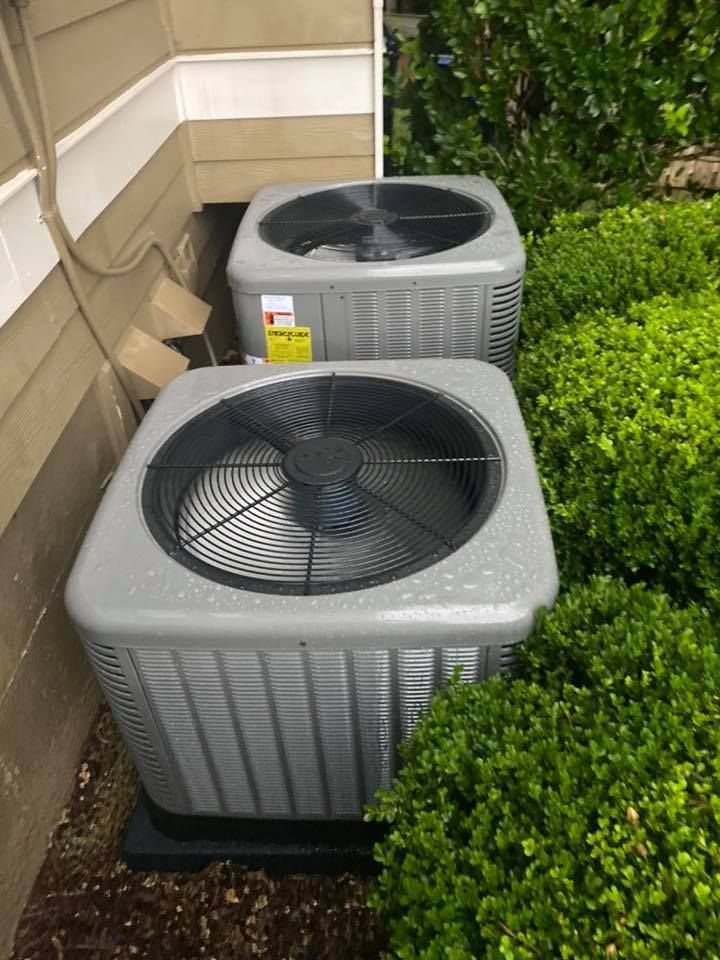 Two outdoor air conditioning units beside a house, near green shrubs and beige siding.
