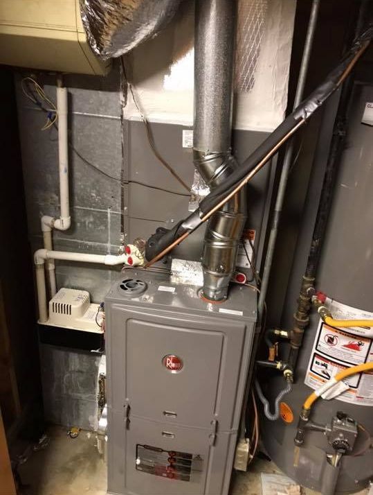 Gray furnace with exposed ducts and pipes in a utility closet, with stacked boxes beside it.