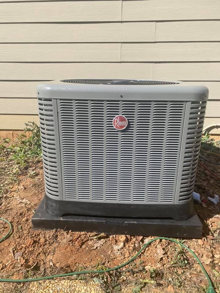 Outdoor air conditioner unit against beige siding with a red logo and dirt around the base