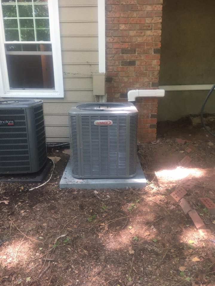 Two outdoor air conditioning units beside a house wall, one on a concrete pad near a brick corner.
