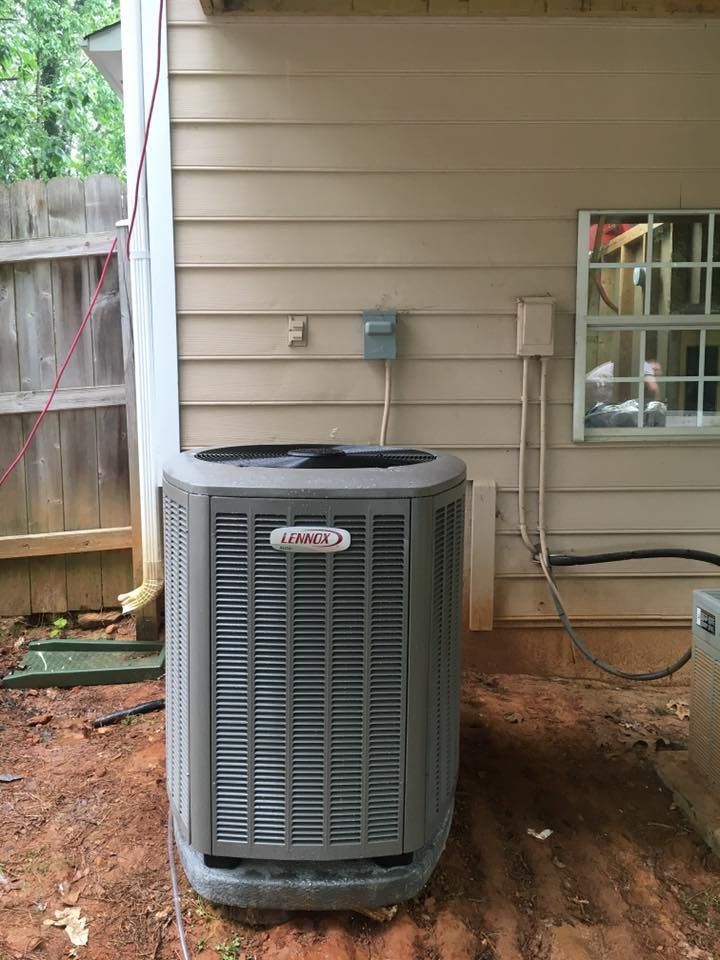 Outdoor air conditioner unit beside a house wall with utility lines and a fenced yard background