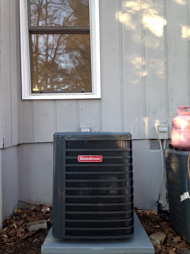 Outdoor air conditioner unit beside a gray house wall under a window, with leaves on the ground
